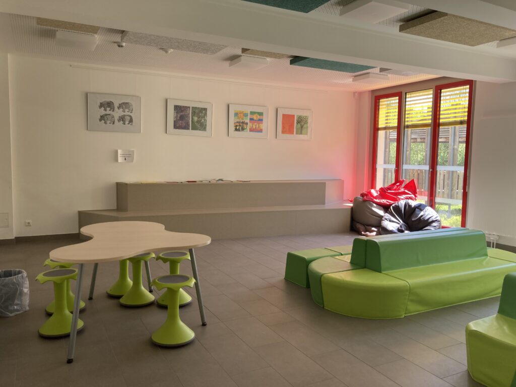 Sitzbereich der Schulbibliothek mit grünen Kunstledersofas und Tisch mit grünen Hockern. An der Wand gibt es Stufen zum Sitzen