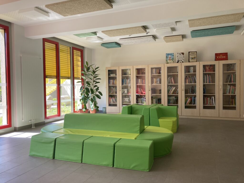 Regalwand mit Büchern. Davor grüne Kunstledersofas und Sessel.